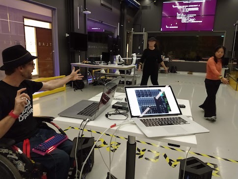 Happy performer in wheelchair. Two computers on a table. Two dancers in the background. A screen with computer code on the back wall.